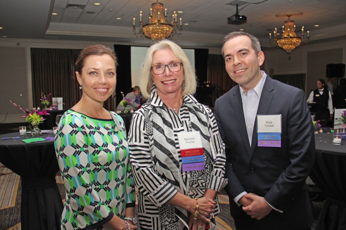 Three people standing in a large room with chandeliers hanging from the ceiling. The person on the left is a woman wearing a green and black patterned dress the person in the middle is a middle-aged woman with blonde hair and glasses and the person to the right is a man wearing a black suit and a purple name tag. They are all smiling and appear to be posing for the photo. In the background there are tables with black tablecloths and flower arrangements.