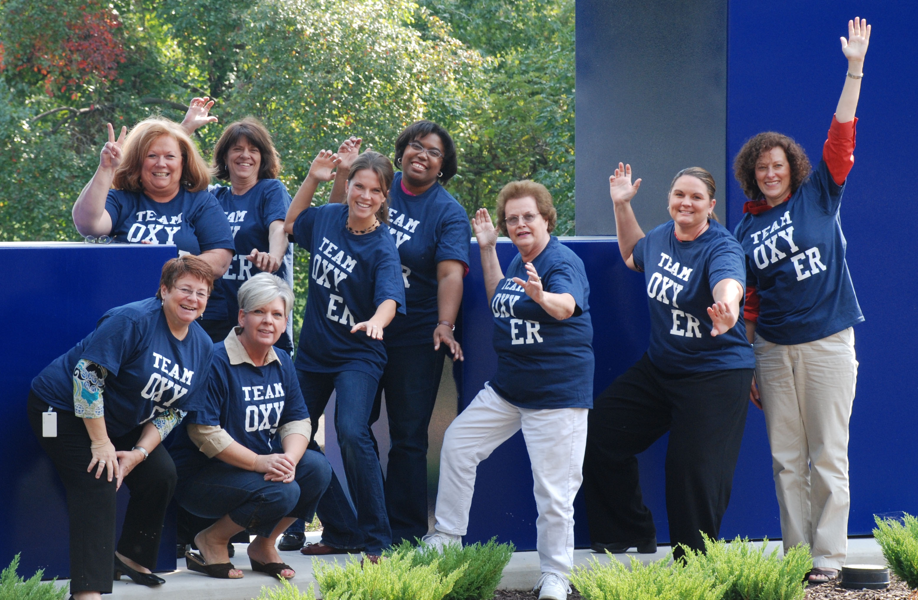 A group of nine women wearing blue t-shirts with the words "Team Oxygen" printed on them. They are standing in front of a blue wall with trees and bushes in the background. The women are posing for a group photo with their hands up in the air making a peace sign with their fingers. They appear to be happy and confident.