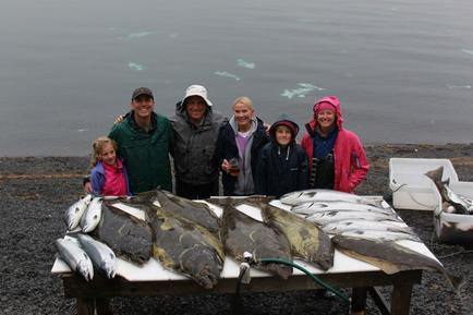 A group of seven people standing around a table with a large number of fish on it. The table is on a rocky beach with the ocean in the background. The people are smiling and posing for the photo. On the left side of the table there is a man wearing a green jacket and a hat and on the right side there are two women wearing pink jackets and hats. In the center of the group there appears to be a man holding a glass of beer. The fish on the table are large and silver and they are arranged in a neat pile. There are also several smaller fish scattered around the table. The sky is overcast and the water is calm suggesting that the photo was taken on a cloudy day.
