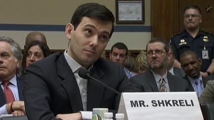 A man sitting at a table with a nameplate that reads "Mr. shebeli". He is wearing a dark suit and tie and appears to be speaking into a microphone. Behind him there are several other people sitting at the table some of whom are looking at him attentively. On the right side of the image there is a police officer standing in front of the table. The background is a room with a blue wall and a wooden panel.