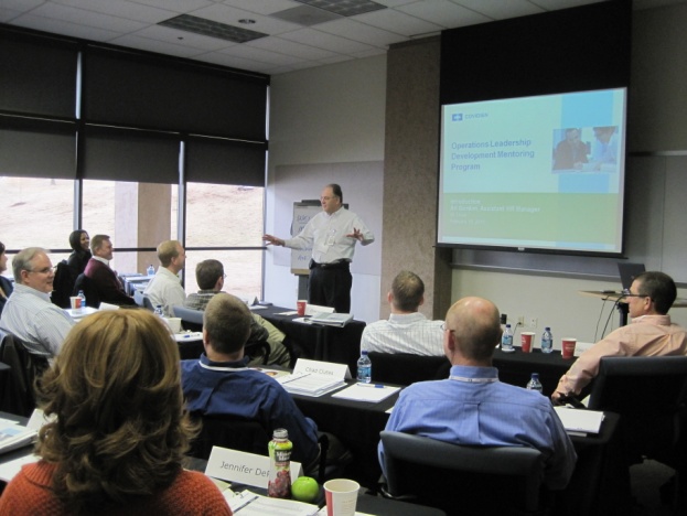 A man giving a presentation to a group of people in a conference room. The room has a large window on the left side and a projector screen on the right side. The man is standing at the front of the room and is giving a speech. He is wearing a white shirt and black pants and is gesturing with his hands as he speaks. The people in the room are sitting at long tables with papers water bottles and nameplates on them. They appear to be listening attentively to the presentation.