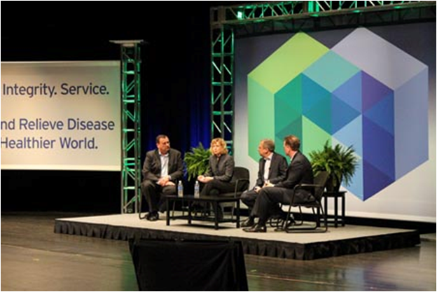 A panel discussion on a stage. There are four people sitting on the stage three men and one woman. They are all dressed in formal business attire and appear to be engaged in a discussion. The stage is decorated with a large screen on the right side and a banner on the left side that reads "Integrity Service and Relieve Disease Healthier World." There are also two potted plants on either side of the stage. The background is dark.