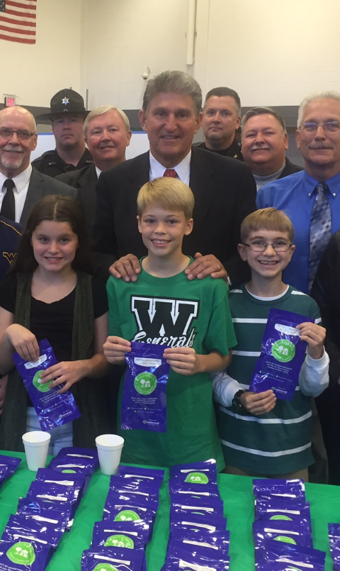 A group of people standing around a table with a green tablecloth. On the table there are several rows of purple packets with the letter "W" on them. The packets are arranged in a neat and orderly manner. In the center of the image there is a young boy wearing a green t-shirt with the letter "W." He is standing with his arms around a man wearing a suit and tie. Behind him there appears to be a man in a police uniform and a woman in a black dress. The people around the table are smiling and looking at the camera. There is an American flag hanging on the wall in the background.