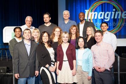 A group of people standing on a stage with a blue curtain in the background. There is a large screen behind them with the words "Imagine the possibilities" written in colorful letters. The people in the photo are of different ages genders and ethnicity's. They are all smiling and appear to be posing for a group photo. Some of them are wearing casual clothes while others are dressed in formal attire. The overall mood of the photo is happy and confident.