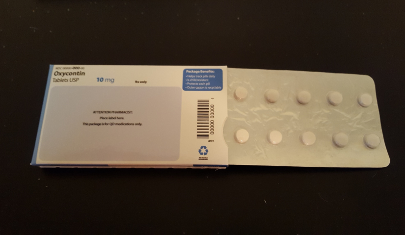 A blister pack of Oxycontin tablets. The blister pack is white in color and has a label on the top left corner that reads "Oxycontin Tablets USP 10 mg". The label also has a barcode and a blue recycling symbol on the bottom right corner. On the right side of the blister pack there are 12 white pills arranged in a circular pattern. The pills appear to be round and have a smooth texture. The pack is lying on a black surface.