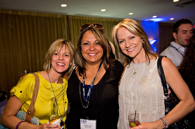 Three women standing together and posing for a photo. They are all smiling and appear to be at an event. The woman on the left is wearing a yellow top and has blonde hair while the woman in the middle has brunette hair and is wearing sunglasses on her head. The third woman is in the center wearing a black top and is holding a drink in her hand. All three women are wearing name tags around their necks. In the background there are other people and a stage with blue lights.