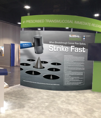 Of a trade show booth with a large banner on the right side. The banner has a black background with white text that reads "1 Prescribed Transmucosal Immediate-Release" and "When Breakthrough Cancer Pain Spikes Strike Fast". On the left side of the banner there is an illustration of a surgical instrument which appears to be a scalpel with a blue handle and a black head. Below the instrument there are six circular holes which are likely used for breaking through cancer pain spikes. The text on the banner is in white and is in a larger font size than the rest of the text. The booth is located in an indoor space with a concrete floor and a green wall in the background.