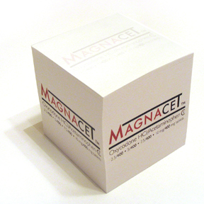 Of a white cube-shaped box with the word "MAGNACET" written in red and black letters on the top and bottom of the box. The box appears to be made of cardboard and has a glossy finish. The word "Magnacet" is written in a bold sans-serif font and is centered on the box with the letters "M" and "C" in a larger font size than the rest of the text. Below the word there is a smaller text that reads "Oxycodone HCL/Acetaminophen" in black letters. The background is plain white making the box stand out.