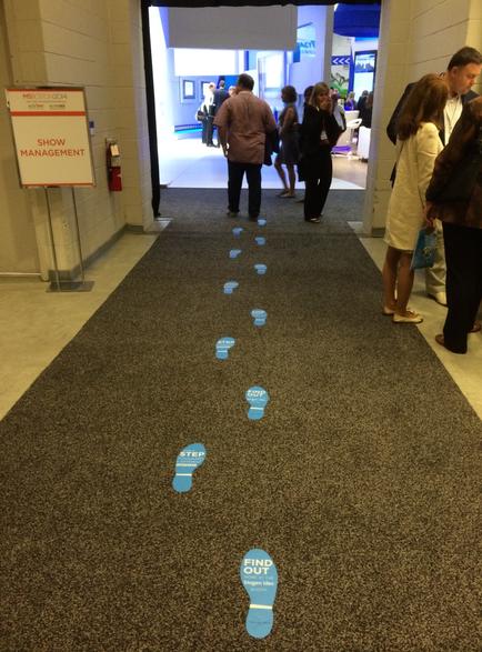 A long carpeted hallway with a group of people walking on it. The carpet is black and has blue footprints scattered across it. On the left side of the image there is a sign that reads "Show Management" and on the right side there are several people standing in line some of them are looking at the sign. The hallway appears to be in an office or conference room as there are other people visible in the background.