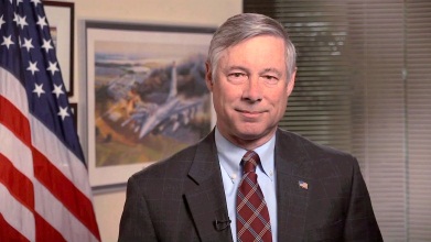 A portrait of a middle-aged man in a suit and tie. He is standing in an office with an American flag on the left side of the image and a painting hanging on the wall on the right side. The man appears to be in his late 60s or early 70s with gray hair and a slight smile on his face. He has a small American flag pin on his lapel. The background is blurred but it seems to be a window with blinds.