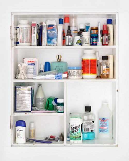 A white medicine cabinet with three shelves. The cabinet is open and the contents are neatly arranged on the shelves. On the top shelf there are several bottles of different sizes and colors. The bottom shelf has a few bottles of products including a blue bottle of deodorant a green bottle of lotion and a white bottle of soap. The background is plain white.