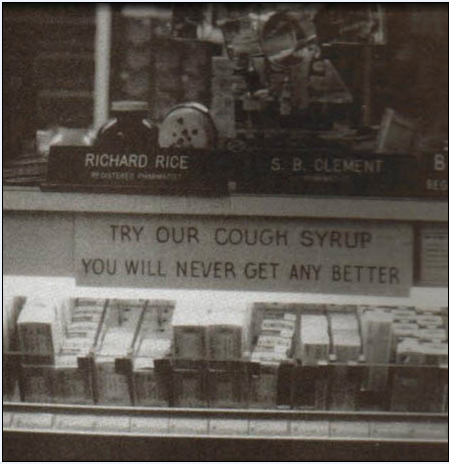 A black and white photograph of a display case in a store. The display case is filled with various items such as a sign that reads "Richard Rice S.B. Clement Try Our Cough Syrup You Will Never Get Any Better". The sign is written in bold white letters on a black background. Below the sign there is a smaller sign that says "Try our cough syrup" in smaller white letters. On the right side of the display case there are several boxes of cough syrup stacked on top of each other. The boxes appear to be of different sizes and colors. The background is blurred but it appears to be a store window with other items visible.