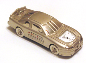 A photograph of a miniature model of a vintage car. The car is made of metal and has a shiny metallic finish. It has a sleek streamlined design with a pointed nose and a curved roofline. The body of the car is painted in a light beige color with the word "MAGNACET" written in red and white on the side. The front of the body has a large clock face with black numbers and hands indicating the time. The wheels are silver and appear to be made of rubber. The model is sitting on a white surface and the background is blurred making the car the focal point of the image.