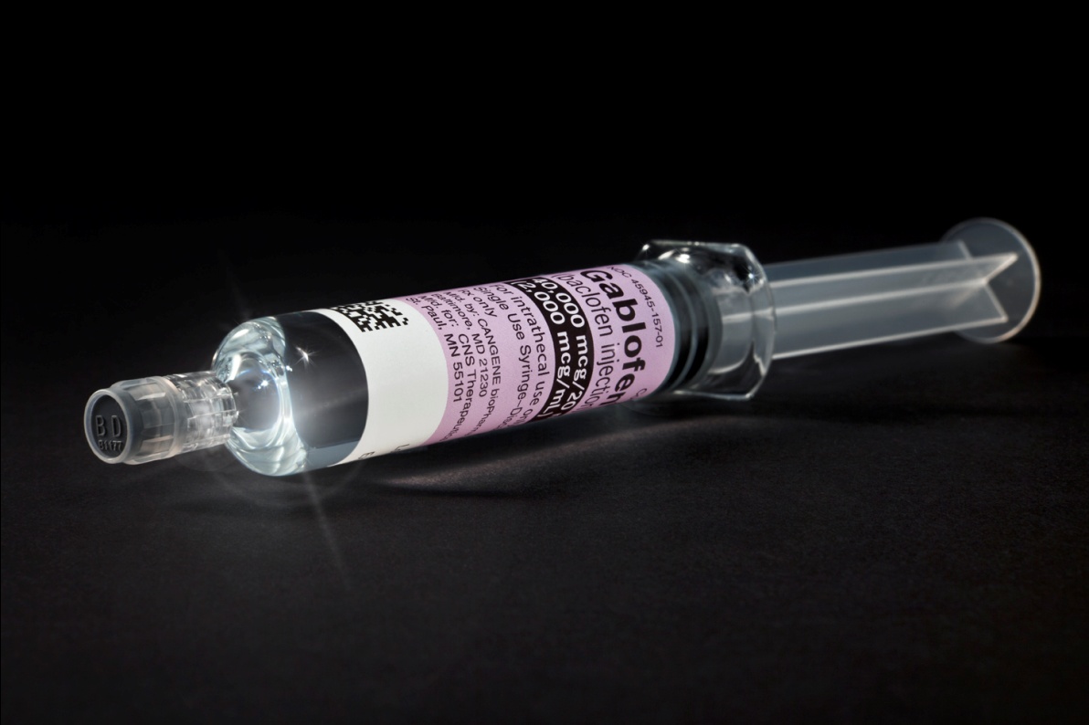 A syringe lying on a black background. The syringe is made of clear plastic and has a long cylindrical body with a transparent cap on one end. The cap is slightly tilted to the side and the syringe appears to be empty. On the side of the cap there is a label with a pink and white design. The label has some text written on it but it is not clear what it says. The background is completely black making the syringes stand out.