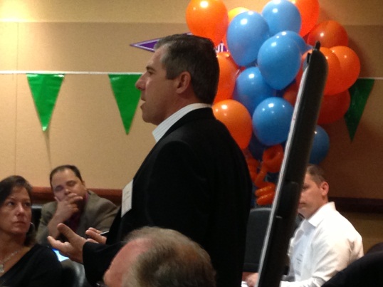 A man standing in front of a group of people in a conference room. He is wearing a black suit and appears to be speaking into a microphone. Behind him there is a wall decorated with colorful balloons in shades of orange blue and green. The people in the room are sitting in chairs and appear to be listening attentively to the man speak.