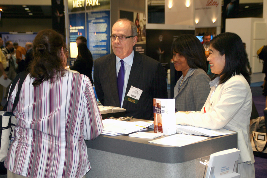 A group of people at an exhibition or trade show. There are three people in the image two women and one man standing behind a reception desk. The man is wearing a suit and tie and appears to be speaking to the two women. They appear to be engaged in a conversation. In the background there are other people walking around and a banner that reads "Meet Pain."