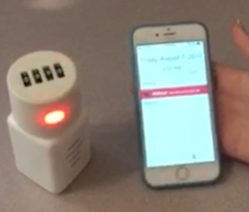 A person's hand holding an iPhone with a white device next to it. The device appears to be a small cylindrical device with a red light on top. The phone screen displays a message that reads "Friday August 7 2015". The background is blurred but it seems like the person is standing on a countertop.