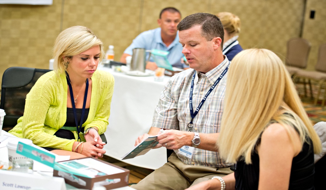 A group of people sitting around a table in a conference room. There are three people in the image two women and one man. The woman on the left is blonde wearing a yellow cardigan is holding a pen and appears to be explaining something to the man on the right. The man in the middle is a man wearing a plaid shirt and is looking at a piece of paper in his hand. He is also wearing a blue lanyard around his neck. The table is covered with a white tablecloth and there are nameplates and cups on it. In the background there are other people sitting at the table and a projector screen on the wall.