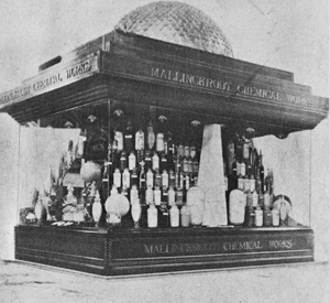 A black and white photograph of a display case with a dome on top. The display case is made of wood and has a rectangular shape with a flat roof. The top of the display case has a sign that reads "Mallinckrodt Chemical Works".<br /><br />Inside the case there are several bottles of different sizes and shapes arranged in a neat and orderly manner. There are also some decorative items such as a vase a lamp and a statue of a bird.<br /><br />The display case appears to be made of a dark wood and is mounted on a pedestal. The background is plain and unadorned with no other objects or decorations visible.