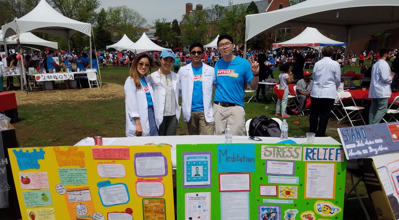 A group of five young people standing in front of a table with a poster on it. The poster has various images and text on it including "Stress Relief" and "Meditation". The people are wearing white lab coats and are smiling at the camera. Behind them there are several white tents and tables with people sitting at them. The sky is blue and there are trees and buildings in the background. It appears to be an outdoor event or gathering.