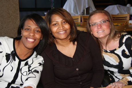 Three women sitting together and smiling at the camera. They appear to be in a restaurant or cafe as there are tables and chairs visible in the background.