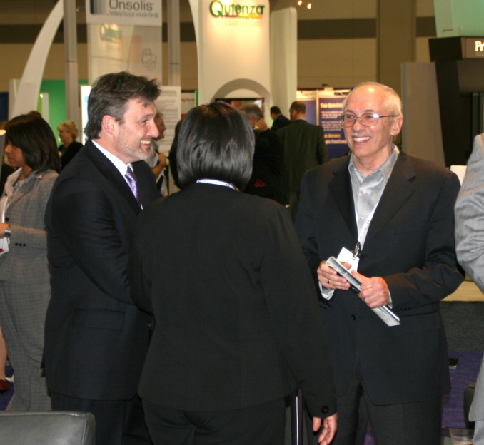 Three people two men and a woman engaged in a conversation at an exhibition. They are all dressed in formal business attire with the man on the left wearing a black suit and tie the man in the middle wearing a gray suit and glasses and the woman on the right wearing a white blouse and black blazer. All three people are smiling and appear to be engaged in the conversation. In the background there are other people walking around and a booth with a sign that reads "Onsolis" and "Quenza".