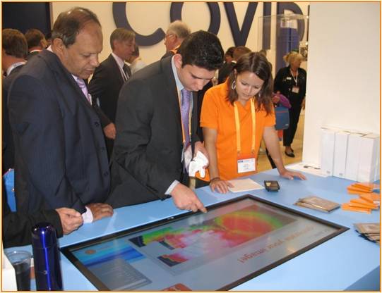 Three people two men and a woman standing around a table with a large screen in front of them. The screen displays a colorful image of a map with different colors and shapes. The man on the left is wearing a suit and tie the man in the middle is wearing an orange shirt and the woman on the right is holding a piece of paper. They appear to be looking at the screen intently. There are other people in the background but they are not visible. The table is covered with a blue tablecloth and there are orange folders and a water bottle on the table. It appears to be an exhibition or trade show booth.