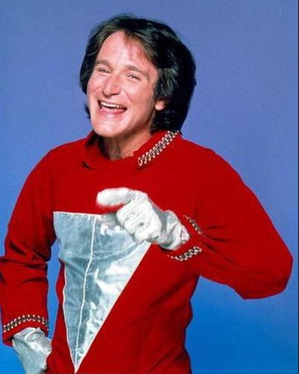 A portrait of a man who appears to be in his late 60s or early 70s. He is wearing a red jacket with a silver corset-like design on the front. The jacket has a high collar and long sleeves. He has a pair of white gloves on his hands and is smiling widely. The background is a solid blue color. The man has shoulder-length dark hair and is looking directly at the camera with a slight smile on his face.