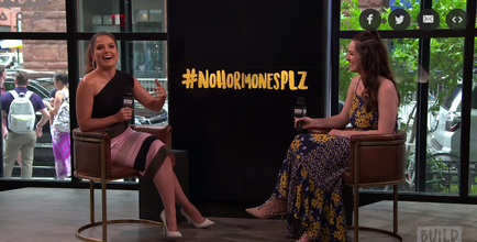 Two women sitting in chairs facing each other and engaged in a conversation. The woman on the left is wearing a black one-shoulder dress with a pink skirt and white heels. She is holding a microphone and appears to be speaking to the other woman. The background shows a large window with a view of a city street and a building. On the right side of the image there is a sign that reads "#NOMORMONESPLZ" in yellow letters. Both women are smiling and seem to be having a good time.