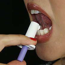 A close-up of a person's mouth with their mouth open and their tongue sticking out. The person is holding a small white and purple spray bottle that looks like a medical device. The bottle appears to be being used to administer a medication under the person's tongue. The background is black and the focus is on the person and the spray bottle.