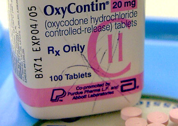 A white pill bottle with a pink label that reads "OxyContin 20 mg (oxycodone hydrochloride controlled-release) tablets". The label also has the Rx Only logo and the words "100 Tablets" written on it. The bottle is sitting on a blue surface and there are a few pink pills scattered around it.