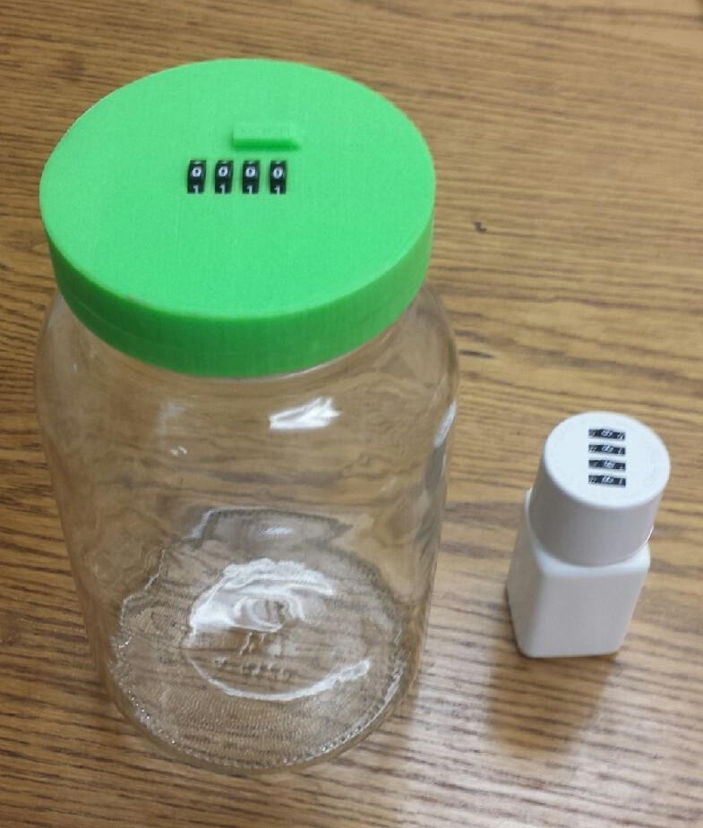 A small glass jar with a green lid. The jar appears to be empty and is sitting on a wooden surface. Next to the jar there is a small white salt shaker with a black and white label on it. The lid of the jar has a green button on it.