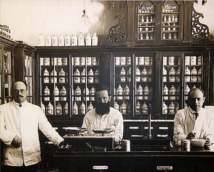 A black and white photograph of three men standing behind a desk in a laboratory. The desk is made of wood and has a large mirror above it. The men are wearing lab coats and appear to be in their late twenties or early thirties. On the left side of the desk there is a man wearing a white lab coat and glasses. He is standing with his hands in his pockets and is looking directly at the camera. On the right side there are two men sitting at the desk. The man in the middle has a beard and is wearing a lab coat. He has a serious expression on his face and is holding a pen in his right hand. The other two men are sitting behind the desk and are looking at the man on the left with a slight smile on their faces. The background of the image shows a large wooden cabinet with multiple shelves filled with bottles and jars. The cabinet appears to be ornate and ornate with intricate carvings on the top and sides.