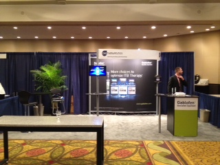 A conference room with a blue curtain in the background. In the center of the room there is a large white banner with the logo of a company called "Gazelle" on it. The banner also has an image of a computer screen and text that reads "More choice to experience ITB therapy". On the right side of the banner there are two tables with a man standing behind them speaking into a microphone. On the left side there appears to be a desk with a computer monitor and a plant. The room has a carpeted floor and a chandelier hanging from the ceiling.