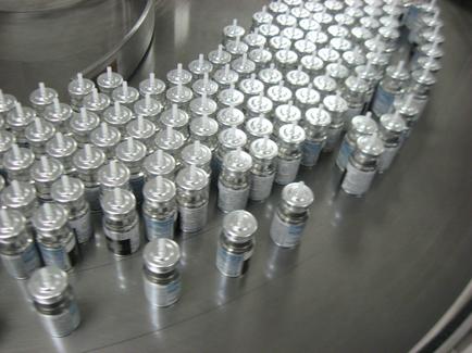 A large number of small metal containers arranged in a circular pattern on a conveyor belt. The containers are of different sizes and shapes with some being round and others being cylindrical. They are all silver in color and have a label on the front with a blue and white design. The belt appears to be made of stainless steel and has a shiny surface. The background is blurred but it seems to be a factory or industrial setting.