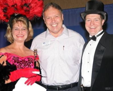 Three people posing for a photo. On the left is a woman wearing a pink and black dress with a large red feathered headpiece. She is holding a white purse and is smiling at the camera. In the middle is a middle-aged man wearing a striped shirt and black pants. He is also smiling and appears to be in a good mood.<br /><br />On the right is a young man in a black tuxedo and top hat. He has a big smile on his face. The background is a blue curtain.