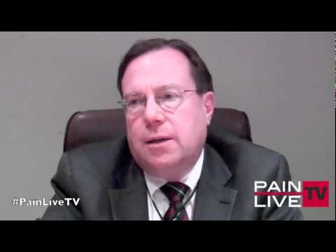 A screenshot from a video interview. It shows a man in a suit and tie sitting in an office chair. He appears to be in his late 40s or early 50s with short dark hair and glasses. He is looking directly at the camera with a serious expression on his face. The background is a plain white wall. On the bottom right corner of the image there is a logo for PainLiveTV.