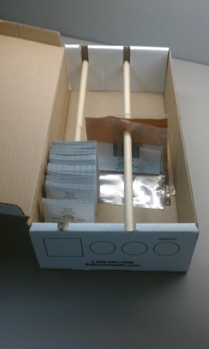 A small open cardboard box with a fold-open lid and two wooden dowels across the top. Pieces of paper with holes punched through them hang from the left dowel. On the right plastic sleeves with holes in them hang from the dowel. The background is a plain white surface.