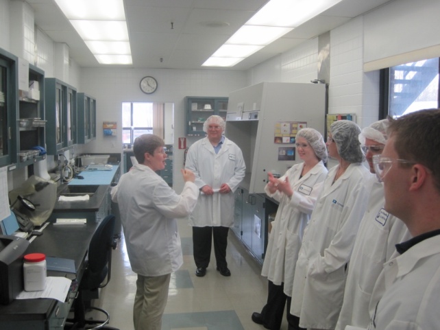 A group of six people in a laboratory setting. They are all wearing white lab coats and are standing in front of a large machine. The machine appears to be a control panel or a control center. There are several shelves and cabinets on the left side of the image and a desk on the right side. The people in the image are engaged in a conversation with one person gesturing with their hands as if they are explaining something to the others. The room has a high ceiling with recessed lighting and a clock on the wall.