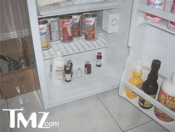 An open refrigerator with the door open. Inside the refrigerator there are several cans of food and bottles of condiments. On the left side of the image there is a cardboard box with some items inside. The refrigerator appears to be in a room with a tiled floor and a white wall in the background. The door is open revealing the contents inside.