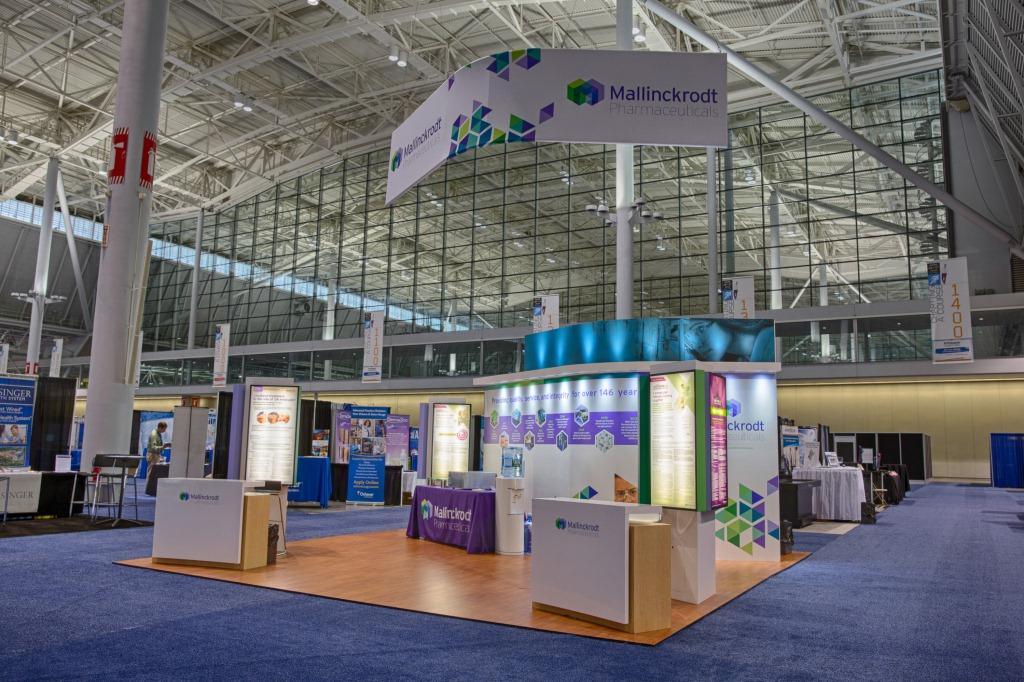 A large exhibition hall with a high ceiling and large windows. The hall is decorated with a blue carpet and white walls. In the center of the hall there is a large white booth with a colorful design and a sign that reads "Mallinckrodt". The booth is surrounded by other booths and tables. There are also banners and signs hanging from the ceiling. The overall atmosphere of the exhibition hall is modern and professional.