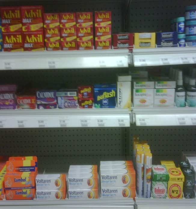 A shelf in a grocery store filled with various types of food items. On the left side of the shelf there are several boxes of Advil arranged in rows. The boxes are red and yellow in color and have the brand name "Advil" written on them. Next to them there is a carton of Lumbar and a pack of Voltaren. <br /><br />On the right side on the bottom right there appears to be a variety of other products including bottles of toothpaste toothbrushes and toothpaste tubes. There are also a few other items on the shelf. The shelf is white and has a grid-like pattern.