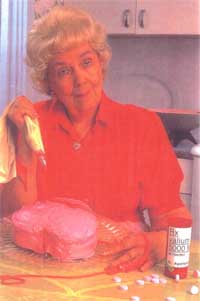 An elderly woman sitting at a kitchen counter with a large pink cake in front of her. She is wearing a red polo shirt and has blonde curly hair. The woman is holding a yellow spatula and appears to be in the process of decorating the cake. There are several pills scattered around the counter and a bottle of pills on the right side of the image. The background shows a kitchen with white cabinets and a window with curtains.