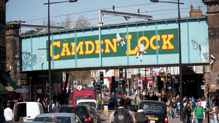 A busy street scene in London UK. The focal point of the image is a large blue bridge with the words "Camden Lock" written in yellow and red letters. Above the bridge there is a mural of two people jumping in the air. The bridge appears to be over a busy intersection with cars buses and pedestrians. The street is lined with brick buildings on both sides and there are traffic lights and street lamps visible. The sky is blue and the overall atmosphere is busy and bustling.